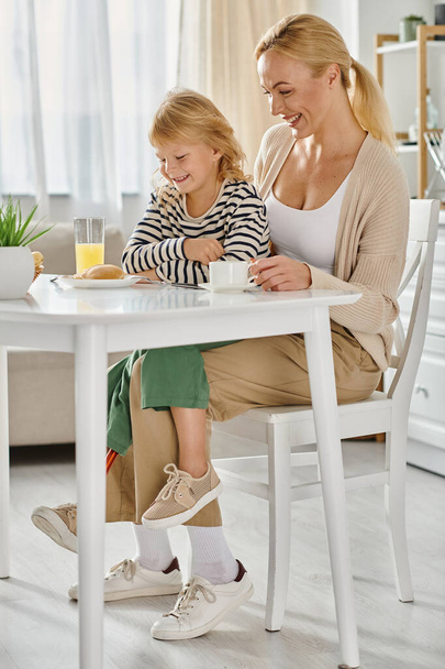 child with prosthetic leg sitting on laps of happy mother and looking at pancakes during breakfast - Photo, Image