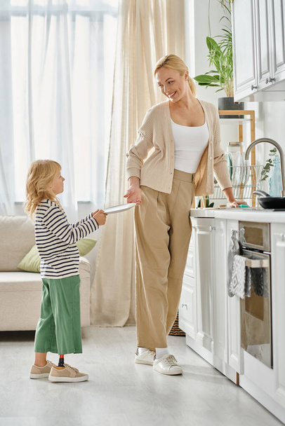χαριτωμένο κορίτσι με προσθετικό πόδι κρατώντας πιάτο και βοηθώντας ευτυχισμένη μητέρα πλύσιμο πιάτων στην κουζίνα - Φωτογραφία, εικόνα