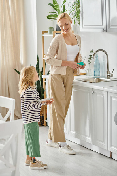 Protez bacaklı küçük kız tabağı tutuyor ve mutlu annenin mutfakta bulaşık yıkamasına yardım ediyor. - Fotoğraf, Görsel
