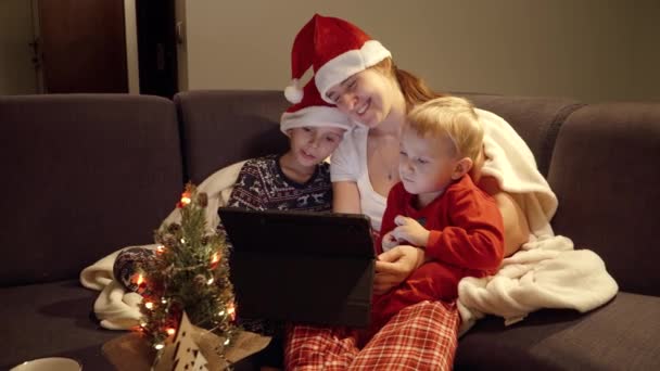 Niños felices con mamá viendo películas en la noche de Navidad. Familia usando tableta. Vacaciones de invierno, celebraciones y fiestas - Metraje, vídeo