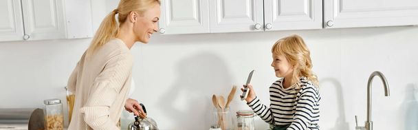 boldog kislány ül a pulton, és mutatja okostelefon anya főzés a konyhában, banner - Fotó, kép