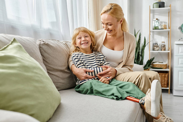 šťastná matka objímající dceru s protézou nohy a sedí spolu na gauči v obývacím pokoji - Fotografie, Obrázek