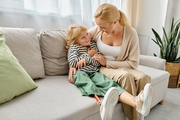 fröhliche Mutter umarmt Tochter mit Beinprothese und sitzt zusammen auf Couch im Wohnzimmer - Foto, Bild