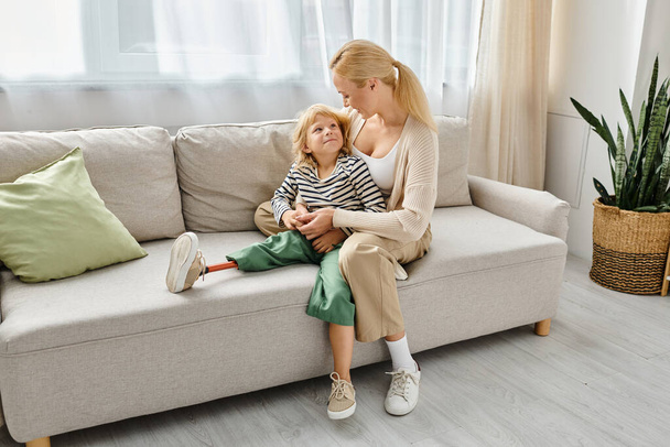 μητέρα αγκαλιάζει ευτυχισμένη κόρη με προσθετικό πόδι και κάθεται μαζί στον καναπέ στο σαλόνι - Φωτογραφία, εικόνα