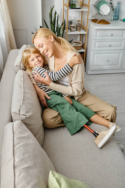 cheerful mother hugging daughter with prosthetic leg and sitting together on sofa in living room - Photo, Image