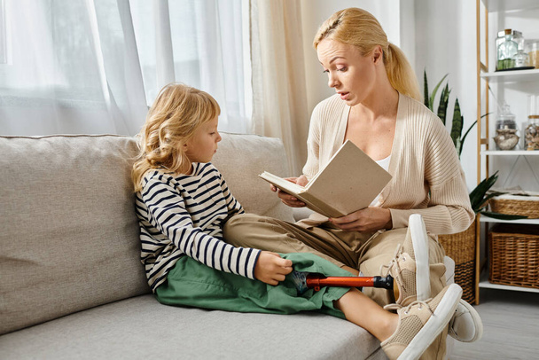 blond vrouw voorlezen boek naar dochter met prothese been terwijl zitten samen in woonkamer - Foto, afbeelding