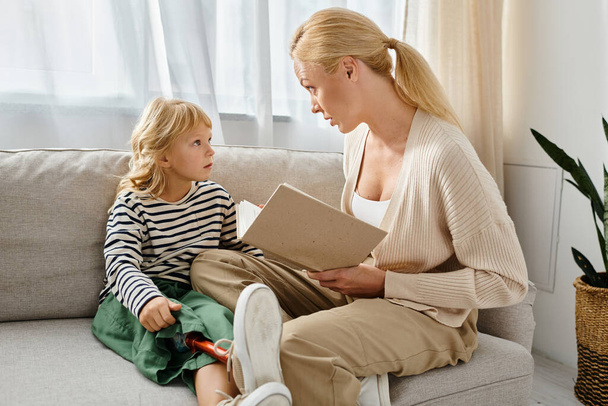 blonde mother reading book to little girl with prosthetic leg while sitting together in living room - Photo, Image