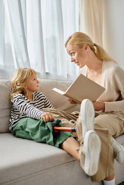 Blonde Mutter liest süßer Tochter mit Beinprothese Buch und sitzt zusammen im Wohnzimmer - Foto, Bild