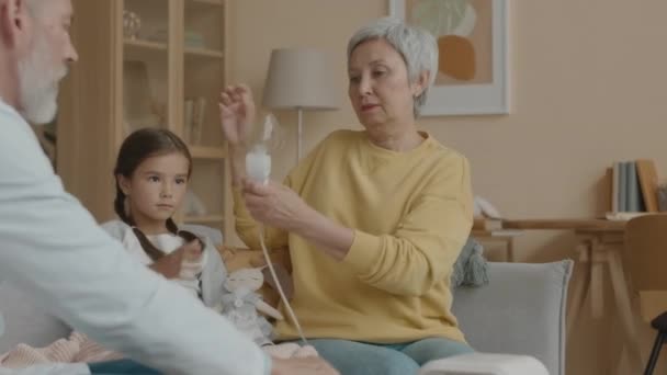 Média tiro de asmático asiático menina usando inalador durante médicos visita em casa sentado com a mãe no sofá em brilhante sala de estar - Filmagem, Vídeo