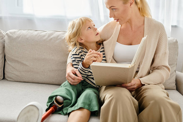 blonde mother reading book to her kid with prosthetic leg while sitting together in living room - Photo, Image