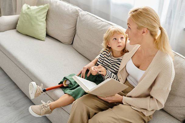 blonde mother reading book to cute child with prosthetic leg while sitting together in living room - Photo, Image