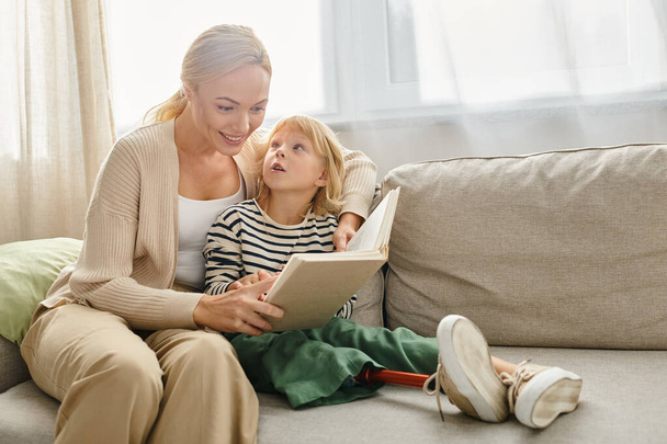 feliz madre leyendo el libro a su hija con la pierna protésica y sentados juntos en la sala de estar - Foto, Imagen