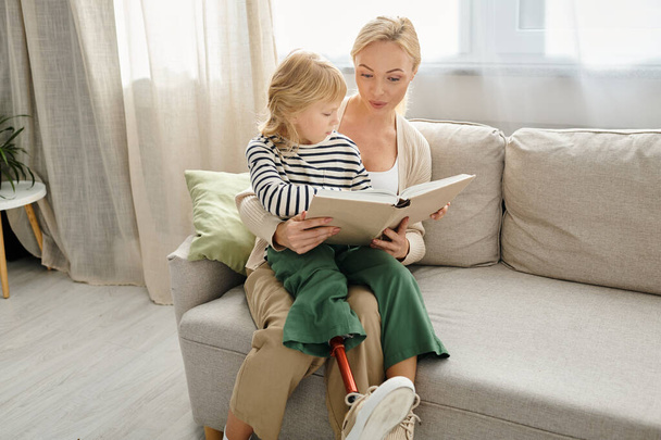 menina feliz com a perna protética sentado em voltas de mãe loira e livro de leitura na sala de estar - Foto, Imagem
