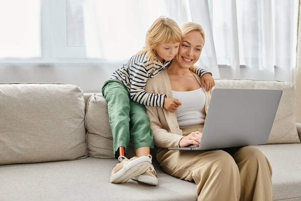 chica feliz con prótesis pierna abrazando a la madre rubia trabajando en el ordenador portátil en la sala de estar moderna - Foto, Imagen