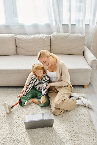 szczęśliwe dziecko z protezą nogi oglądając film na laptopie i siedząc na dywanie razem z matką - Zdjęcie, obraz