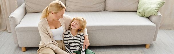 glückliche blonde Frau umarmt fröhliche kleine Tochter in der Nähe der Couch im modernen Wohnzimmer, Banner - Foto, Bild