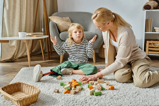 chica confundida con la pierna protésica sentado en la alfombra cerca de juguetes de madera y madre, gesto de encogimiento de hombros - Foto, Imagen