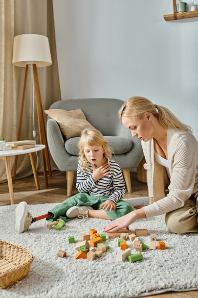 menina confusa com prótese perna sentado no tapete e olhando para brinquedos de madeira perto da mãe - Foto, Imagem