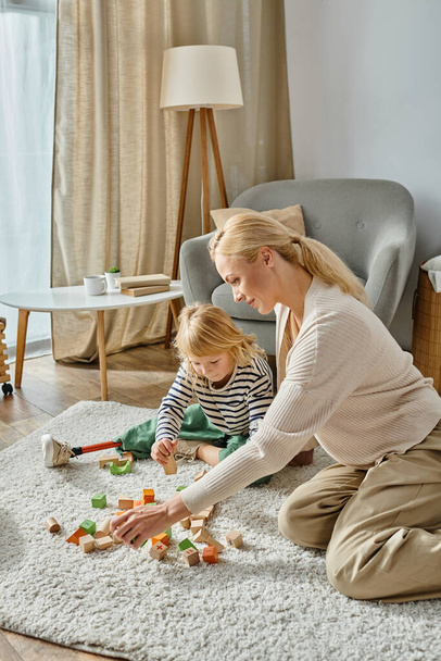 маленька дівчинка з протезною ногою сидить на килимі і грає з дерев'яними іграшками біля матері - Фото, зображення