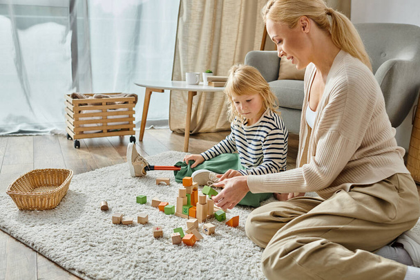 Kleines Mädchen mit Beinprothese sitzt auf Teppich und spielt mit Holzspielzeug neben freudiger Mutter - Foto, Bild