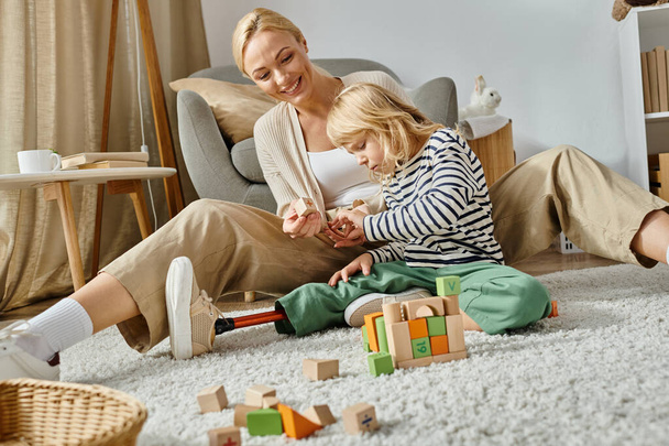 Protez bacaklı küçük bir kız halının üzerinde oturuyor ve neşeli annesinin yanında tahta oyuncaklarla oynuyor. - Fotoğraf, Görsel