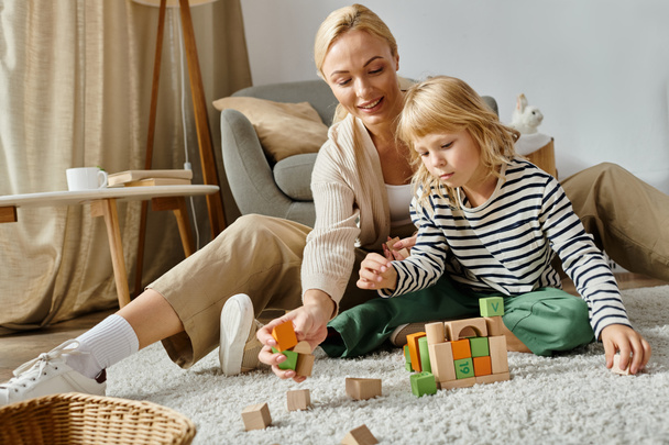kuur meisje met kunstbeen zittend op tapijt en spelen met houten blokken in de buurt van gelukkige moeder - Foto, afbeelding