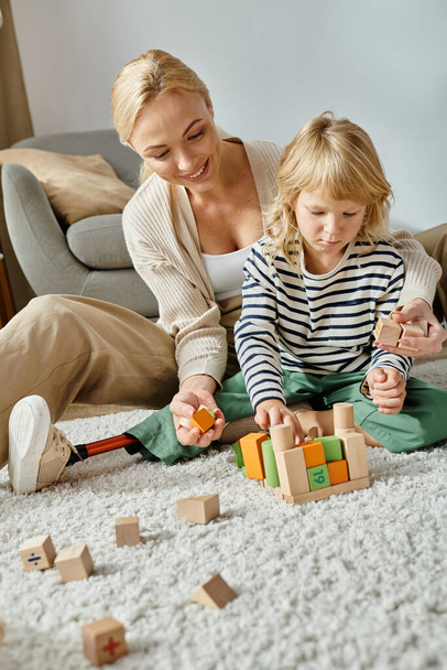 niña con la pierna protésica sentado en la alfombra y jugando con bloques de madera cerca de la madre feliz - Foto, imagen