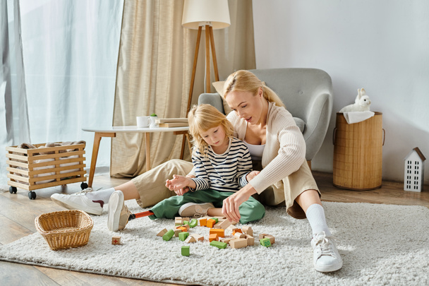 blond meisje met prothese been zitten op tapijt en spelen met houten blokken in de buurt van moeder thuis - Foto, afbeelding