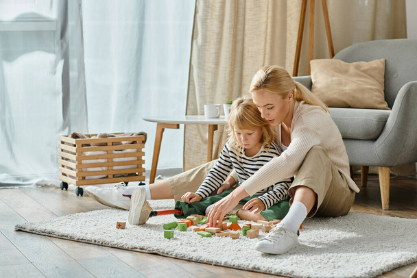 Protez bacaklı sarışın kız halıda oturuyor ve annesinin yanında tahta blok oyunu oynuyor. - Fotoğraf, Görsel