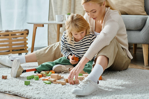 chica rubia con la pierna protésica sentado en la alfombra y jugando con bloques de madera cerca de la madre bonita - Foto, Imagen