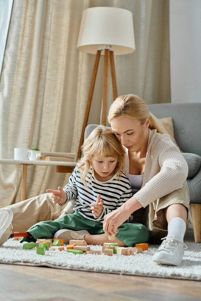 fille blonde avec prothèse jambe assise sur le tapis et jouer avec des blocs de bois près de maman attentionnée - Photo, image