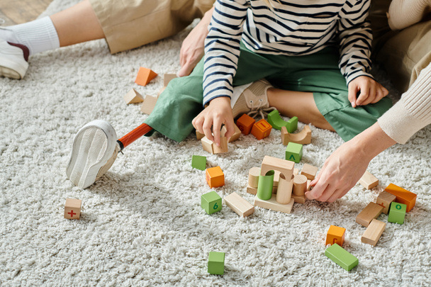 tiro recortado, chica con la pierna protésica sentado en la alfombra y jugando con bloques de madera cerca de la madre - Foto, imagen