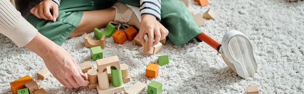 обрізаний банер, дівчина з протезною ногою сидить на килимі і грає з дерев'яними іграшками біля матері - Фото, зображення