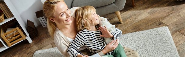 happy woman hugging daughter and looking away while sitting on carpet together, banner - Foto, imagen