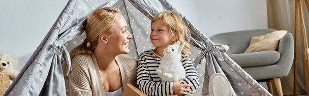 menina bonito segurando brinquedo macio perto de sua mãe alegre leitura livro e sentado em jogar tenda, banner - Foto, Imagem