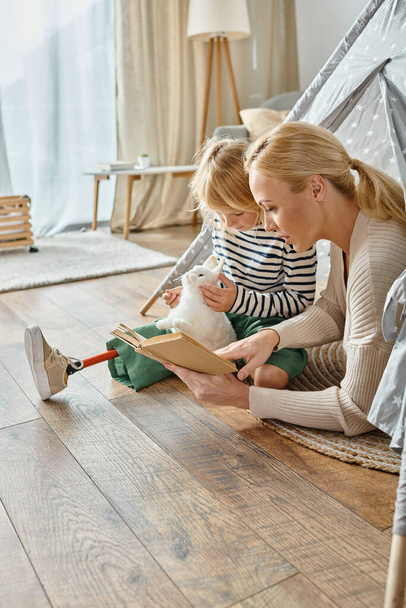 linda chica con prótesis pierna celebración suave juguete y escuchar cómo madre lectura libro en juego tienda de campaña - Foto, imagen