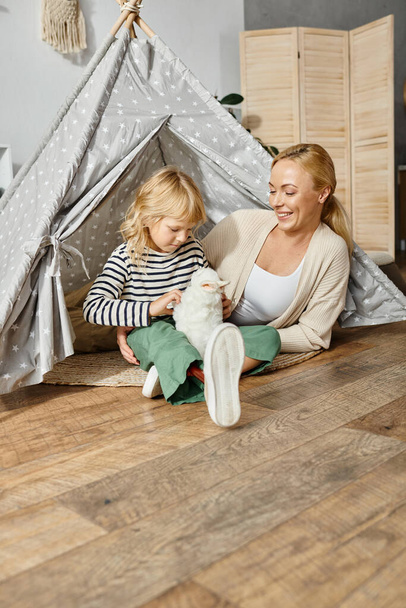 menina loira com prótese perna brincando com brinquedo macio coelho perto de mãe enquanto sentado em jogar tenda - Foto, Imagem