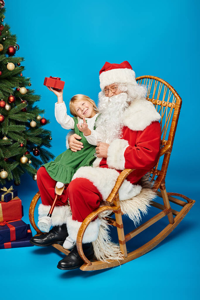 menina feliz com prótese perna sentado em voltas de Papai Noel com caixa de presente ao lado da árvore de Natal - Foto, Imagem