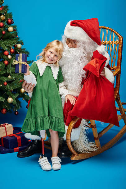 vidám lány műláb gazdaság ajándék közelében Mikulás mellett karácsonyfa kék - Fotó, kép