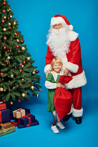 happy girl with prosthetic leg holding sack bag near Santa Claus next to Christmas tree on blue - Photo, Image