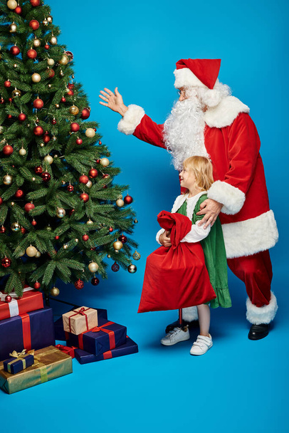 cheerful girl with prosthetic leg holding sack bag near Santa Claus next to Christmas tree on blue - Photo, Image