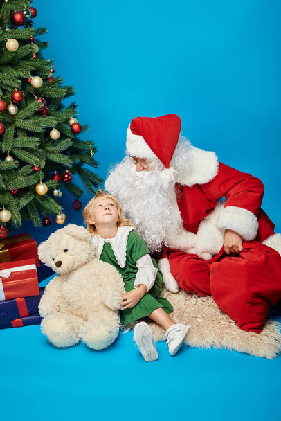 щаслива дитина з протезною ногою і плюшевим ведмедем сидить з Санта Клаусом поруч з ялинкою - Фото, зображення