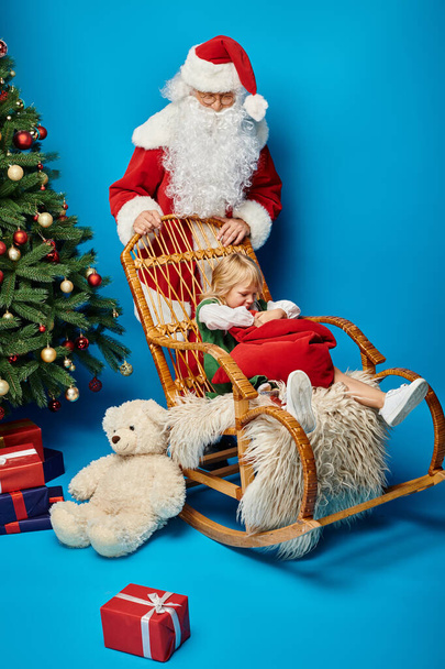 Weihnachtsmann Schaukelstuhl mit süßem Mädchen mit Beinprothese hält Sacksack in der Nähe des Weihnachtsbaums - Foto, Bild