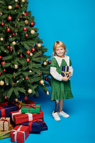 niña rubia con la pierna protésica de pie junto al árbol de Navidad con regalos envueltos - Foto, Imagen