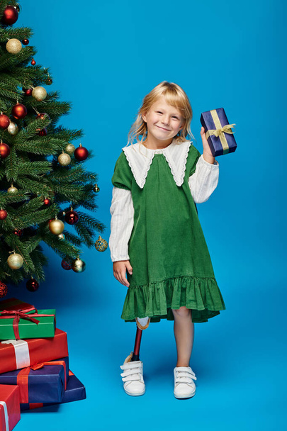 blij klein meisje met kunstbeen houden verpakt cadeau naast kerstboom op blauw - Foto, afbeelding