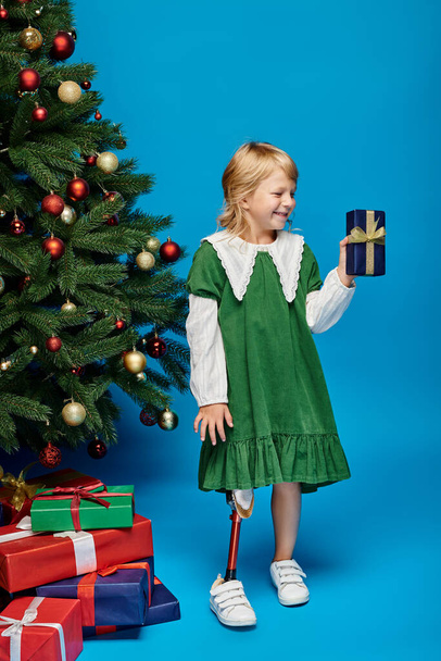 позитивна маленька дівчинка з протезною ногою тримає загорнутий подарунок поруч з ялинкою на синьому - Фото, зображення