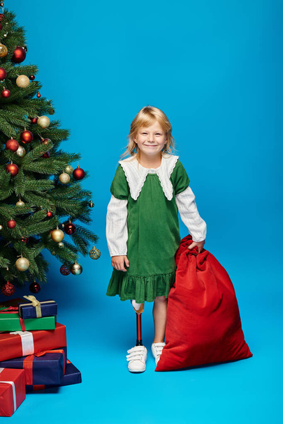 garoto satisfeito no vestido com prótese perna segurando saco saco com presentes perto da árvore de Natal em azul - Foto, Imagem