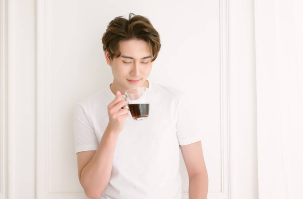 Feliz joven asiático bebiendo una taza de café en una mañana de verano, relajándose con bebida caliente en casa. Milenial coreano o japonés hombre disfrutando de una bebida caliente en fin de semana, tomando un descanso en la sala de estar - Foto, Imagen