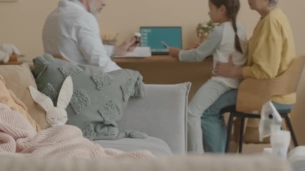 Rabbit toy lying on pillow under soft blanket on couch in living room in focused foreground while male pediatrician speaking to mother of little girl during doctors home visit in blurred background - Footage, Video