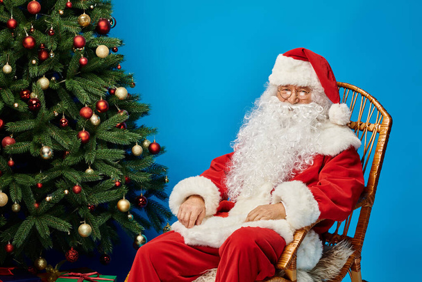 веселый Санта-Клаус с бородой и очками, сидящий в кресле-качалке возле рождественской елки на синей - Фото, изображение
